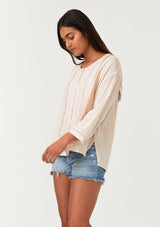 [Color: Light Peach] A half body side facing image of a brunette model wearing a bohemian light pink button front blouse crafted from a linen blend. With long three quarter length sleeves, a round neckline, and delicate pleated details throughout.