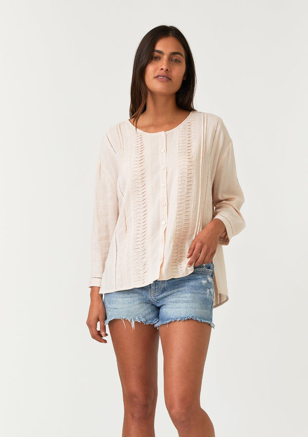 [Color: Light Peach] A half body front facing image of a brunette model wearing a bohemian light pink button front blouse crafted from a linen blend. With long three quarter length sleeves, a round neckline, and delicate pleated details throughout.