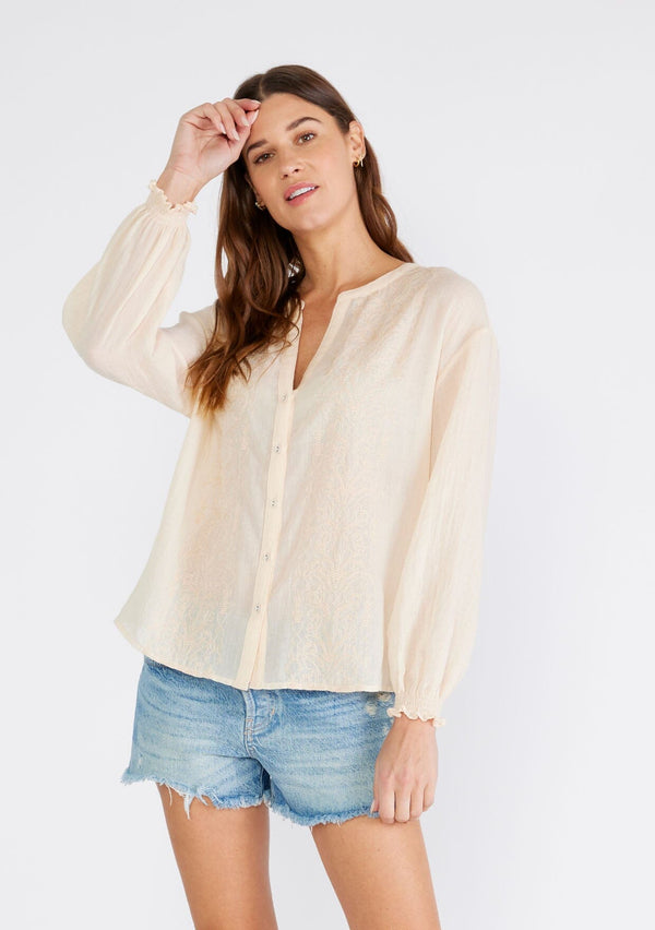 [Color: Light Peach] A front facing image of a brunette model wearing a sheer bohemian blouse in light peach pink. With long sleeves, smocked elastic wrist cuffs, a self covered button front, a split v neckline, and embroidered details along the front. 