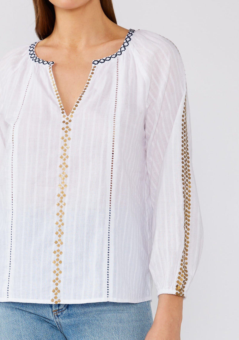 [Color: White/Gold] A close up front facing image of a brunette model wearing a white cotton bohemian blouse with gold sequined details. With long sleeves, a split v neckline, contrast embroidery, and a relaxed fit. 