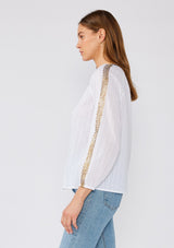 [Color: White/Gold] A side facing image of a brunette model wearing a white cotton bohemian blouse with gold sequined details. With long sleeves, a split v neckline, contrast embroidery, and a relaxed fit. 