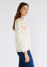 [Color: Vanilla/Coral] A side facing image of a brunette model wearing a beautiful bohemian chic spring blouse in an ivory and coral floral print. With long raglan sleeves, a smocked round neckline, a self covered button front, and a front keyhole. 