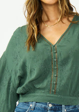 [Color: Forest Green] A detail image of a brunette model wearing a green bohemian top with tonal embroidery, wide sleeves, lattice trims, a button front, and a v neckline. 