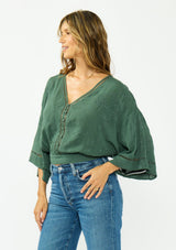 [Color: Forest Green] A side facing image of a brunette model wearing a green bohemian top with tonal embroidery, wide sleeves, lattice trims, a button front, and a v neckline. 