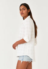 [Color: White] A side facing image of a brunette model wearing a classic bohemian white cotton relaxed shirt in a textured plaid seersucker fabric. With long sleeves, a button tab closure at the sleeve, a collared neckline, and a self covered button front. 