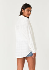[Color: White] A back facing image of a brunette model wearing a classic bohemian white cotton relaxed shirt in a textured plaid seersucker fabric. With long sleeves, a button tab closure at the sleeve, a collared neckline, and a self covered button front. 