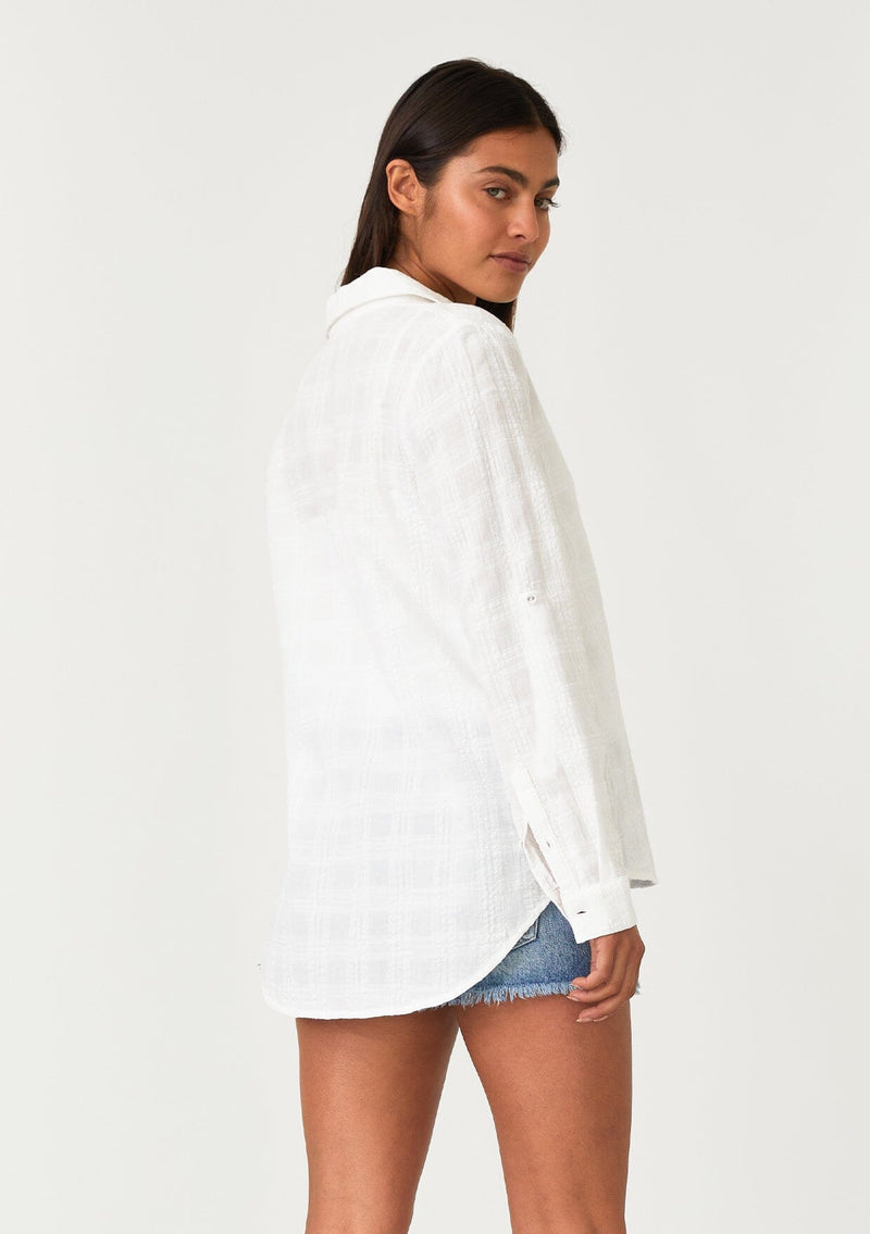 [Color: White] A back facing image of a brunette model wearing a classic bohemian white cotton relaxed shirt in a textured plaid seersucker fabric. With long sleeves, a button tab closure at the sleeve, a collared neckline, and a self covered button front. 