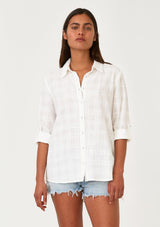 [Color: White] A front facing image of a brunette model wearing a classic bohemian white cotton relaxed shirt in a textured plaid seersucker fabric. With long sleeves, a button tab closure at the sleeve, a collared neckline, and a self covered button front. 