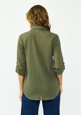 [Color: Olive] A back facing image of a blonde model wearing a green button front shirt crafted in a sheer textured cotton. Featuring a collared neckline, high low hemline, and long roll tab sleeves. Paired with a dark wash denim. 