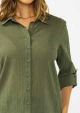 [Color: Olive] A detail image of a blonde model wearing a green button front shirt crafted in a sheer textured cotton. Featuring a collared neckline, high low hemline, and long roll tab sleeves. Paired with a dark wash denim. 