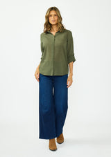 [Color: Olive] A front facing image of a blonde model wearing a green button front shirt crafted in a sheer textured cotton. Featuring a collared neckline, high low hemline, and long roll tab sleeves. Paired with a dark wash denim. 