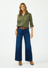[Color: Olive] A front facing image of a blonde model wearing a green button front shirt crafted in a sheer textured cotton. Featuring a collared neckline, high low hemline, and long roll tab sleeves. Paired with a dark wash denim. 
