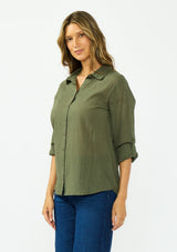 [Color: Olive] A side facing image of a blonde model wearing a green button front shirt crafted in a sheer textured cotton. Featuring a collared neckline, high low hemline, and long roll tab sleeves. Paired with a dark wash denim. 