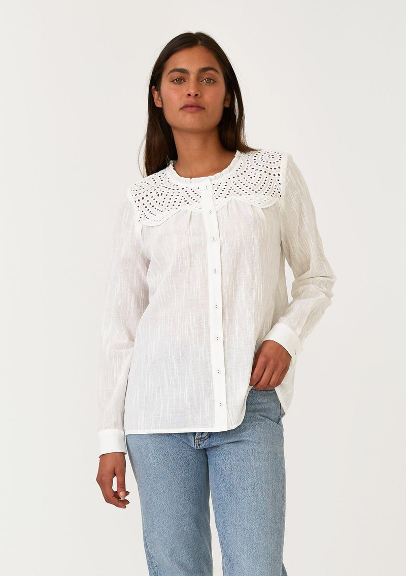 [Color: White] A half body front facing image of a brunette model wearing a white bohemian button front blouse with a ruffled round neckline, a self covered button front, long sleeves, and an eyelet yoke detail. 