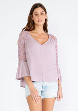 [Color: Dusty Lilac] A half body front facing image of a brunette model wearing a light purple bohemian blouse with a v neckline, a self covered button front, and three quarter length bell sleeves with sheer crochet lace details. 
