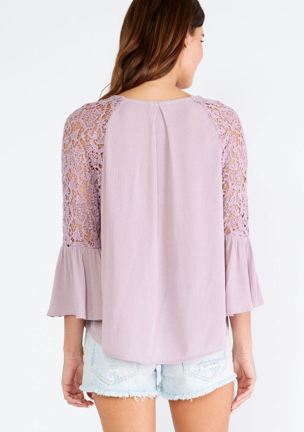[Color: Dusty Lilac] A back facing image of a brunette model wearing a light purple bohemian blouse with a v neckline, a self covered button front, and three quarter length bell sleeves with sheer crochet lace details. 