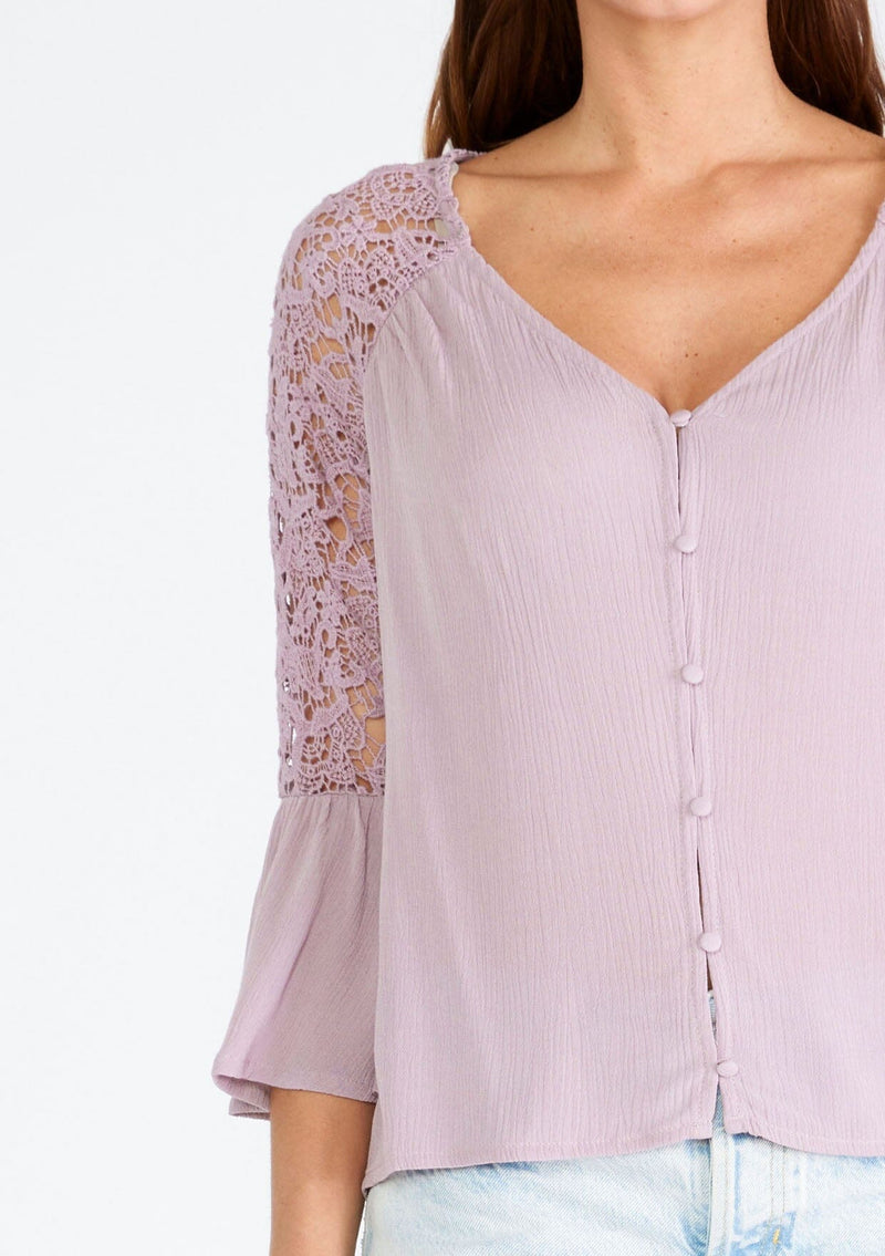 [Color: Dusty Lilac] A close up front facing image of a brunette model wearing a light purple bohemian blouse with a v neckline, a self covered button front, and three quarter length bell sleeves with sheer crochet lace details. 