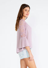 [Color: Dusty Lilac] A side facing image of a brunette model wearing a light purple bohemian blouse with a v neckline, a self covered button front, and three quarter length bell sleeves with sheer crochet lace details. 