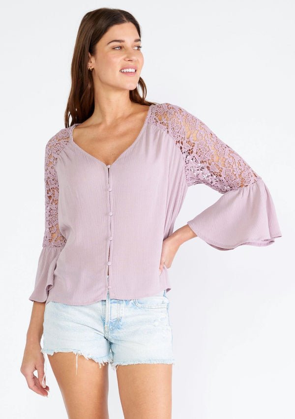 [Color: Dusty Lilac] A front facing image of a brunette model wearing a light purple bohemian blouse with a v neckline, a self covered button front, and three quarter length bell sleeves with sheer crochet lace details. 