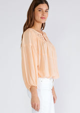[Color: Mellow Peach] A side facing image of a brunette model wearing a peach bohemian blouse with long sleeves, a ruffled elastic wrist cuff, a billowy silhouette, an elastic hemline, and a sheer crochet yoke detail. 