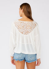 [Color: White] A back facing image of a brunette model wearing a white bohemian blouse with long sleeves, a ruffled elastic wrist cuff, a billowy silhouette, an elastic hemline, and a sheer crochet yoke detail. 