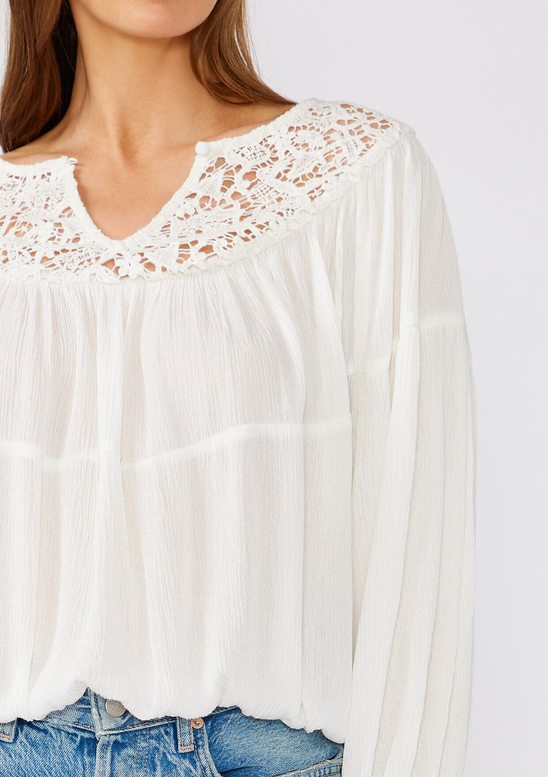 [Color: White] A close up front facing image of a brunette model wearing a white bohemian blouse with long sleeves, a ruffled elastic wrist cuff, a billowy silhouette, an elastic hemline, and a sheer crochet yoke detail. 