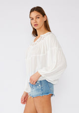 [Color: White] A side facing image of a brunette model wearing a white bohemian blouse with long sleeves, a ruffled elastic wrist cuff, a billowy silhouette, an elastic hemline, and a sheer crochet yoke detail. 