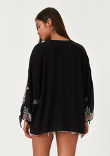 [Color: Black/Rose] A back facing image of a brunette model wearing a bohemian kimono top with a black exterior and pink interior. With voluminous long sleeves, floral embroidery, an open front, and tie wrist cuffs. 