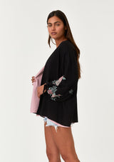 [Color: Black/Rose] A side facing image of a brunette model wearing a bohemian kimono top with a black exterior and pink interior. With voluminous long sleeves, floral embroidery, an open front, and tie wrist cuffs. 