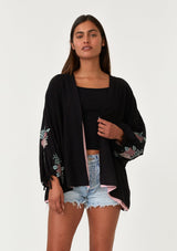 [Color: Black/Rose] A front facing image of a brunette model wearing a bohemian kimono top with a black exterior and pink interior. With voluminous long sleeves, floral embroidery, an open front, and tie wrist cuffs. 