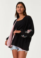 [Color: Black/Rose] A half body front facing image of a brunette model wearing a bohemian kimono top with a black exterior and pink interior. With voluminous long sleeves, floral embroidery, an open front, and tie wrist cuffs. 