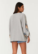 [Color: Silver/Teal] A back facing image of a brunette model wearing a bohemian kimono top with a silver grey exterior and teal blue interior. With voluminous long sleeves, floral embroidery, an open front, and tie wrist cuffs. 