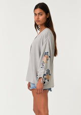 [Color: Silver/Teal] A side facing image of a brunette model wearing a bohemian kimono top with a silver grey exterior and teal blue interior. With voluminous long sleeves, floral embroidery, an open front, and tie wrist cuffs. 