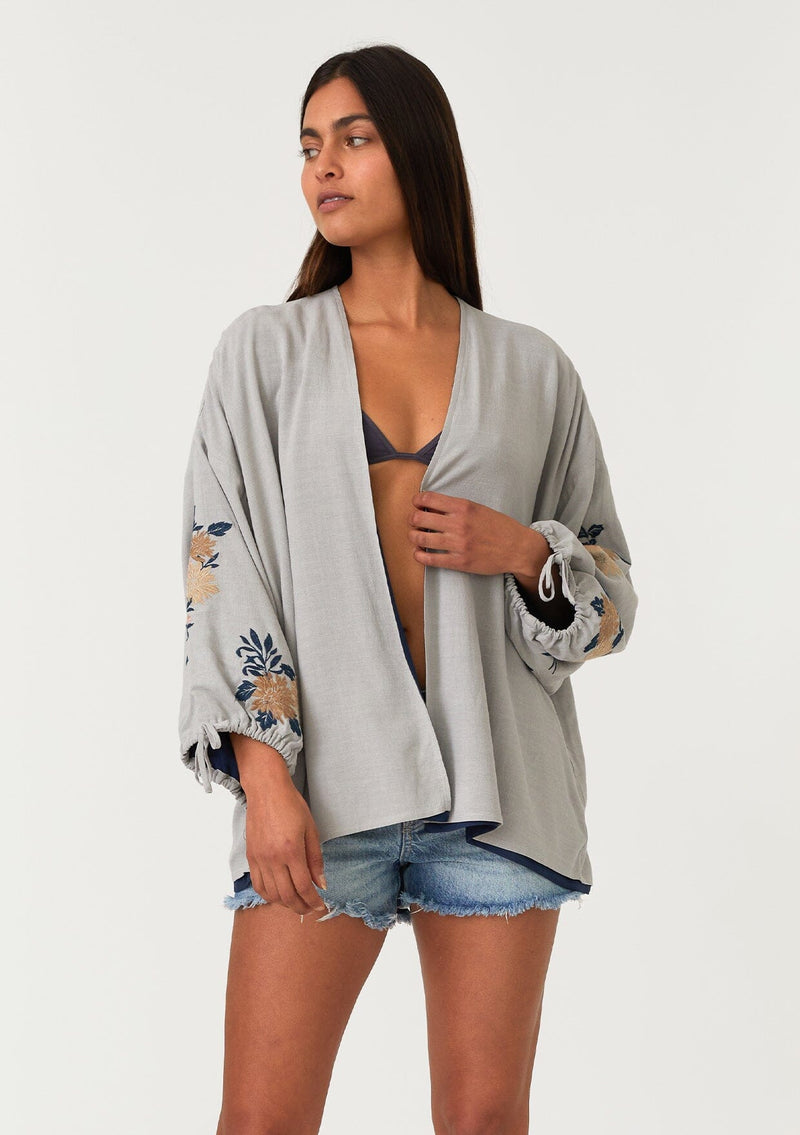 [Color: Silver/Teal] A front facing image of a brunette model wearing a bohemian kimono top with a silver grey exterior and teal blue interior. With voluminous long sleeves, floral embroidery, an open front, and tie wrist cuffs. 