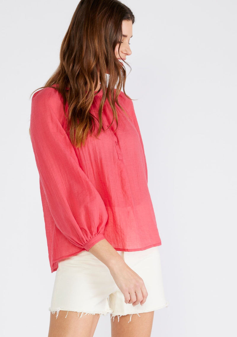 [Color: Coral] A half body side facing image of a brunette model wearing a red sheer bohemian blouse with long raglan sleeves, a round neckline, a button front, and a relaxed fit. 