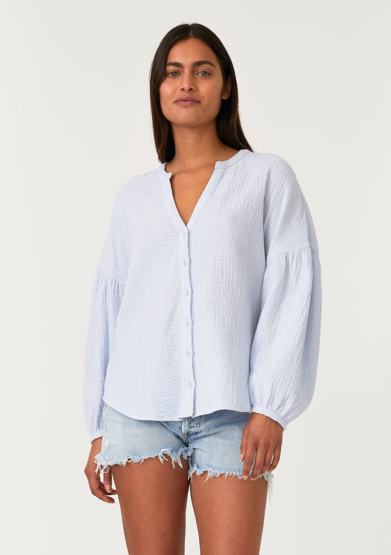[Color: Dusty Blue] A front facing image of a brunette model wearing a classic bohemian blouse in a light blue soft cotton gauze. With voluminous long sleeves, a dropped shoulder, a v neckline, a self covered button front, and a loose, relaxed fit. 