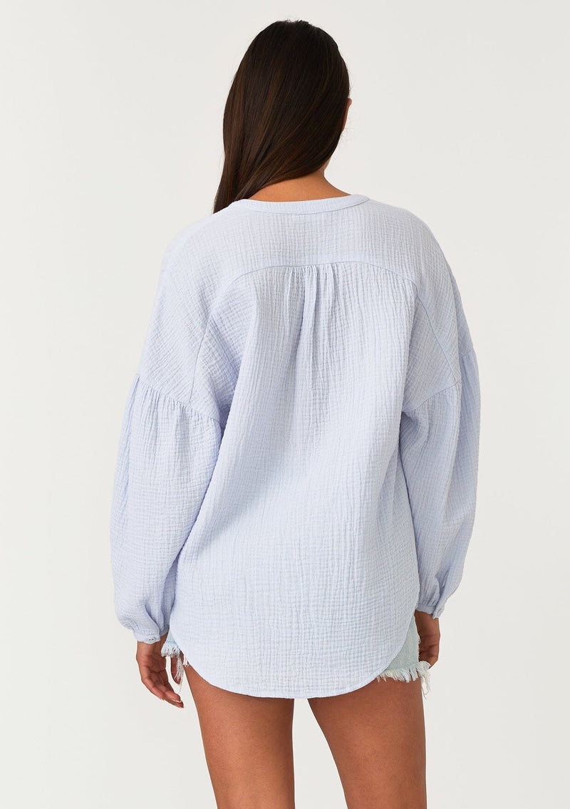 [Color: Dusty Blue] A back facing image of a brunette model wearing a classic bohemian blouse in a light blue soft cotton gauze. With voluminous long sleeves, a dropped shoulder, a v neckline, a self covered button front, and a loose, relaxed fit. 