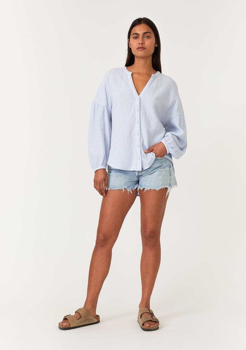 [Color: Dusty Blue] A full body front facing image of a brunette model wearing a classic bohemian blouse in a light blue soft cotton gauze. With voluminous long sleeves, a dropped shoulder, a v neckline, a self covered button front, and a loose, relaxed fit. 