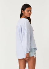 [Color: Dusty Blue] A half body back facing image of a brunette model wearing a classic bohemian blouse in a light blue soft cotton gauze. With voluminous long sleeves, a dropped shoulder, a v neckline, a self covered button front, and a loose, relaxed fit. 
