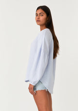 [Color: Dusty Blue] A side facing image of a brunette model wearing a classic bohemian blouse in a light blue soft cotton gauze. With voluminous long sleeves, a dropped shoulder, a v neckline, a self covered button front, and a loose, relaxed fit. 