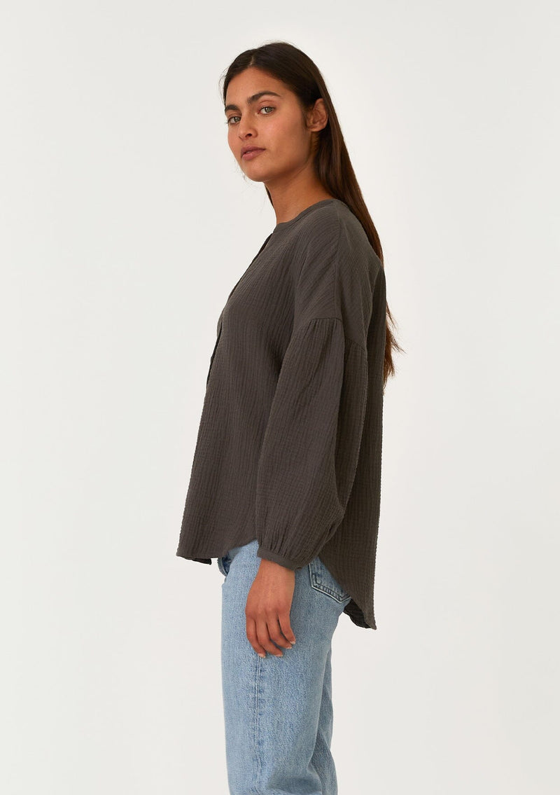 [Color: Lagoon] A side facing image of a brunette model wearing a classic bohemian blouse in an army green soft cotton gauze. With voluminous long sleeves, a dropped shoulder, a v neckline, a self covered button front, and a loose, relaxed fit. 