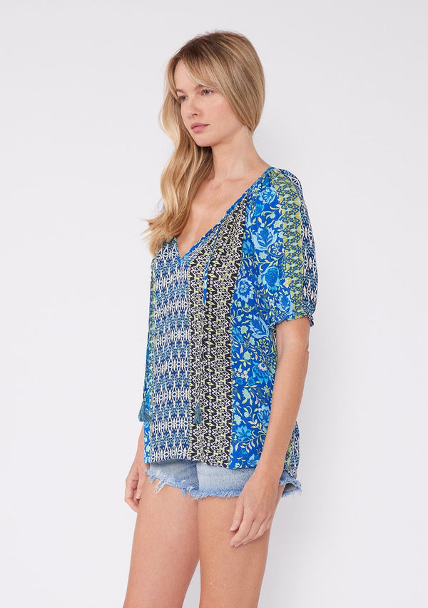 [Color: Blue/Lime] A blonde model wearing an ultra bohemian blouse with a multi print blue floral design. A summer top with a split v neckline, tassel tie front, and short raglan puff sleeves. The perfect summer top for any casual occasion.