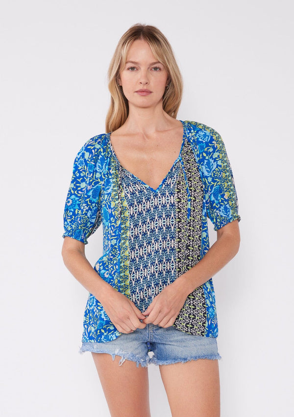 [Color: Blue/Lime] A blonde model wearing an ultra bohemian blouse  with a multi print blue floral design. A summer top with a split v neckline, tassel tie front, and short raglan puff sleeves. The perfect summer top for any casual occasion. 