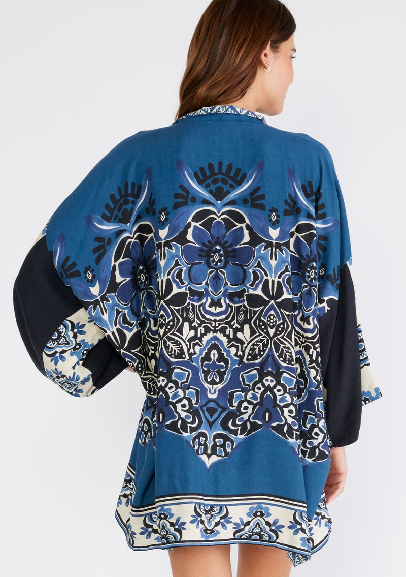 [Color: Indigo/Blue] A back facing image of a brunette model wearing a bohemian kimono in an indigo blue print. With three quarter length long sleeves, an open front, and a mid length. A reversible cover up for the beach or pool side. 