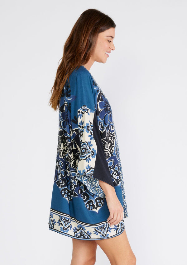 [Color: Indigo/Blue] A side facing image of a brunette model wearing a bohemian kimono in an indigo blue print. With three quarter length long sleeves, an open front, and a mid length. A reversible cover up for the beach or pool side. 