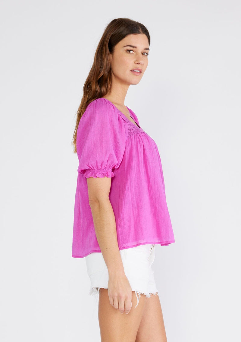 [Color: Orchid] A side facing image of a brunette model wearing a bright purple cotton bohemian top with short puff sleeved, a ruffled elastic cuff, a square neckline, and lace trim. 