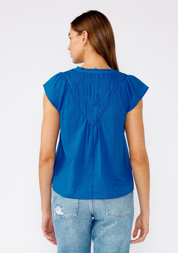 [Color: Cobalt] A back facing image of a brunette model wearing a bright blue bohemian top crafted from lightweight cotton gauze. With short cap sleeves, a split v neckline, gathered details at the yoke, and a raw edge hem. 