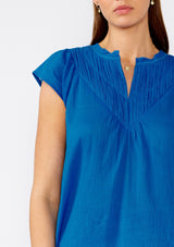 [Color: Cobalt] A close up front facing image of a brunette model wearing a bright blue bohemian top crafted from lightweight cotton gauze. With short cap sleeves, a split v neckline, gathered details at the yoke, and a raw edge hem. 