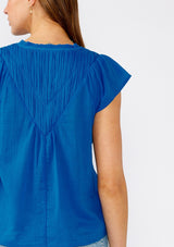 [Color: Cobalt] A close up back facing image of a brunette model wearing a bright blue bohemian top crafted from lightweight cotton gauze. With short cap sleeves, a split v neckline, gathered details at the yoke, and a raw edge hem. 