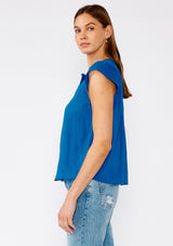 [Color: Cobalt] A side facing image of a brunette model wearing a bright blue bohemian top crafted from lightweight cotton gauze. With short cap sleeves, a split v neckline, gathered details at the yoke, and a raw edge hem. 